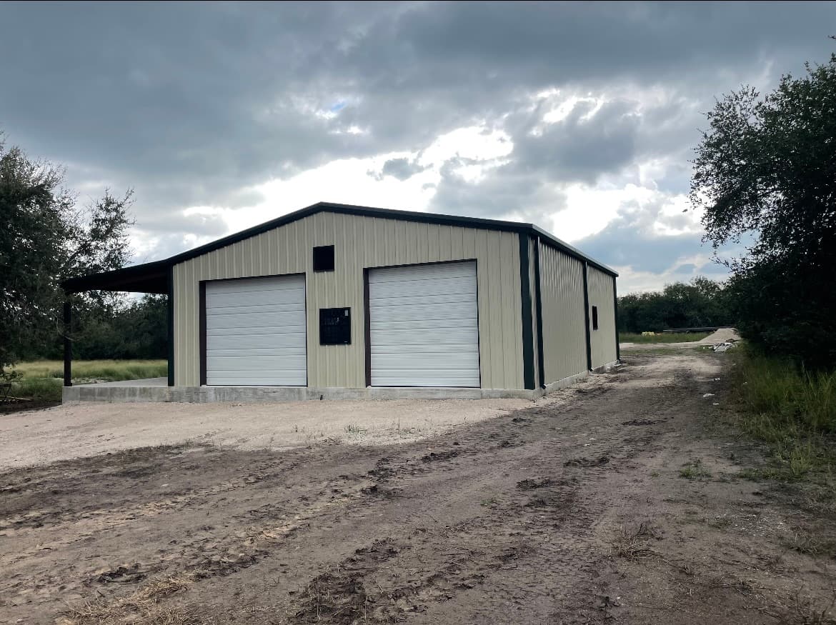 Two-tone cream and forest-green metal shop building with twin roll-up bay doors and an attached open-canopy work area, South Texas