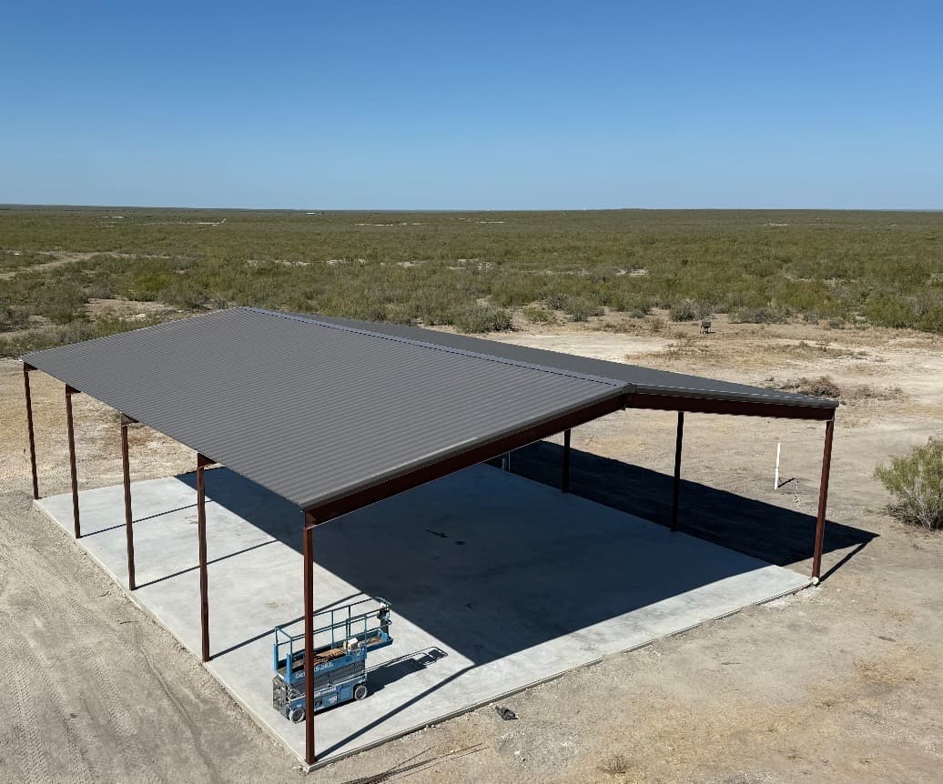 Aerial view of an open-sided equipment cover with charcoal roof and rust-finished columns on a freshly poured slab, South Texas brush country